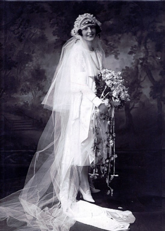 Peggy Fish posando nos anos 20 com um vestido de noiva branco de cetim, véu e buquê de flores. 
