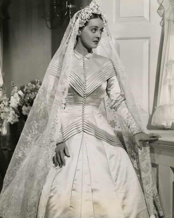Bette Davis em um vestido de noiva branco acetinado e um véu transparente.