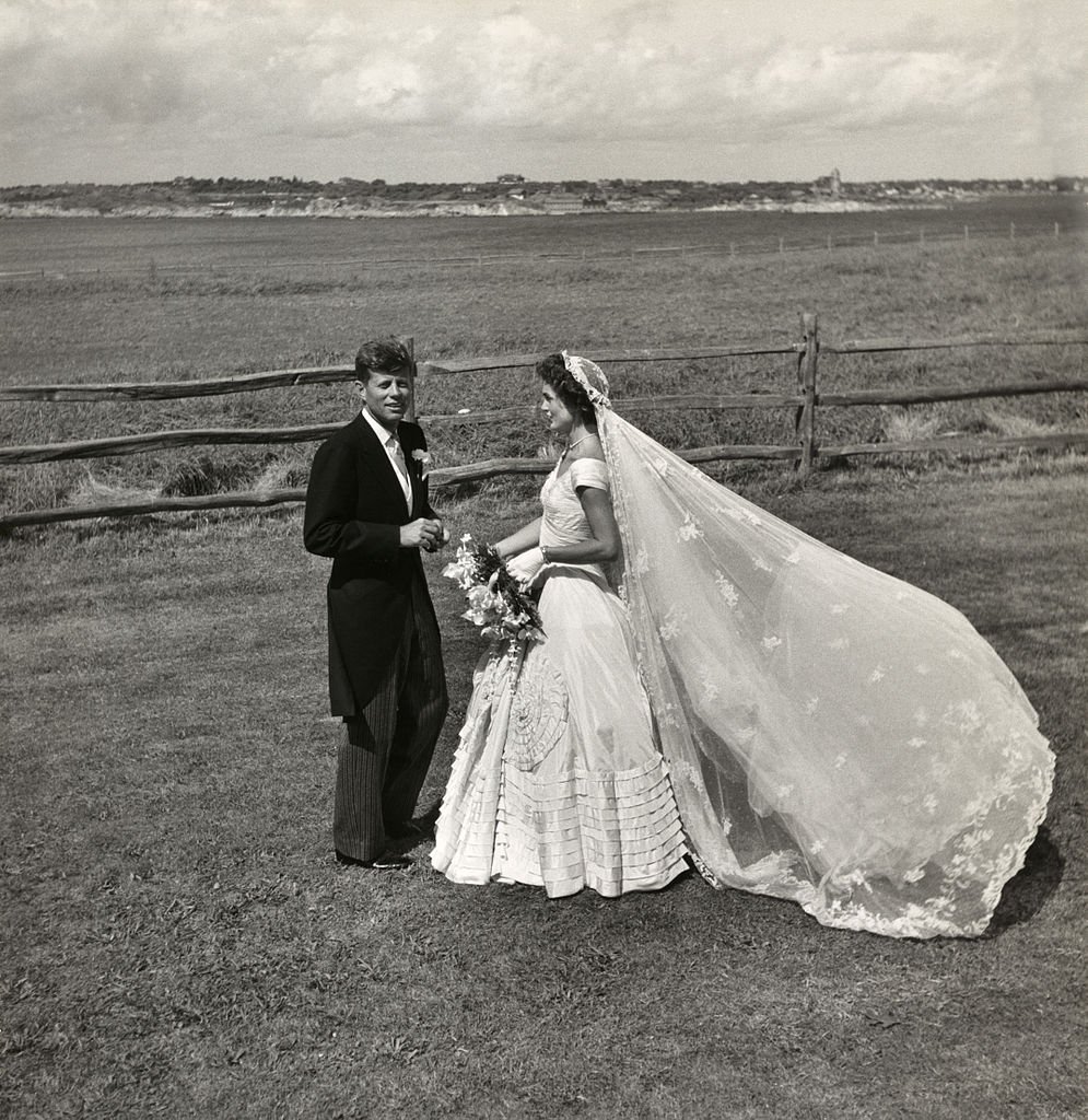 Foto de Jacqueline Kennedy com um vestido de noiva branco e véu transparente ao lado de John Kennedy após o seu casamento, em 1953. 