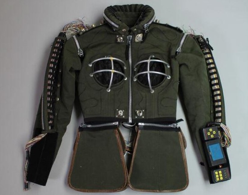 Foto de uma jaqueta verde com diversos tipos de zíper e outros adornos, como um aparelho eletrônico e armações de metal Foto de uma jaqueta verde com diversos tipos de zíper e outros adornos, como um aparelho eletrônico e armações de metal