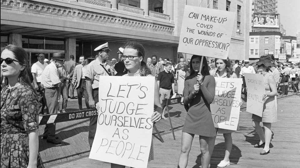Passeata de mulheres em um protesto em 1968 sobre a moda e cidadania contra o concurso Miss America em Nova Jersey, onde se lê "Vamos nos julgar como pessoas" e "Pode a maquiagem disfarças as feridas da opressão?".