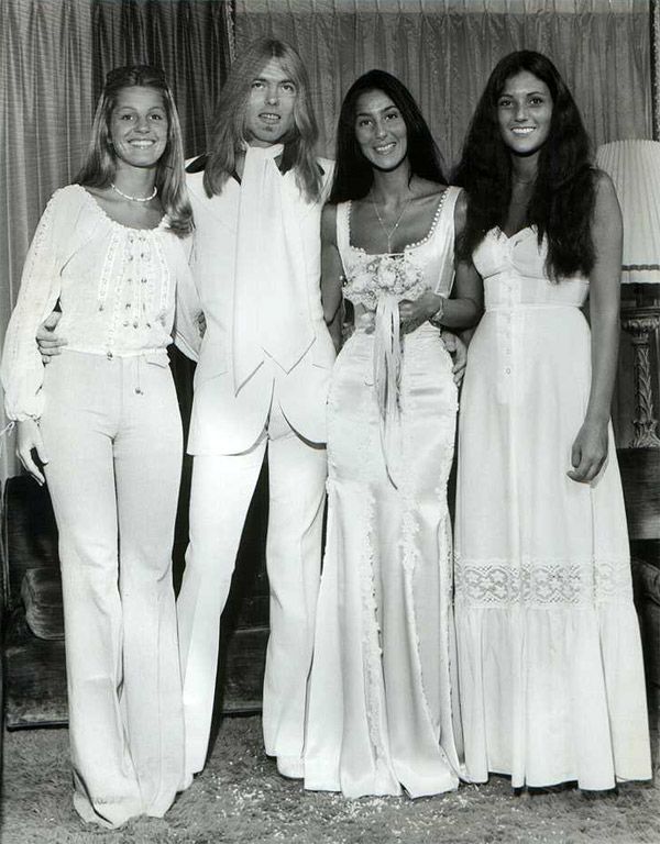 Cher ao lado do seu marido e amigas no seu casamento nos anos 70 com um vestido de noiva branco acetinado e ajustado.