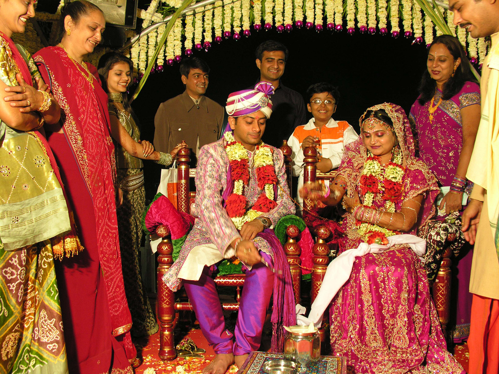 Foto de um casamento hindu com os noivos ao centro vestindo roupas muito coloridas e ricamente ornamentadas. 