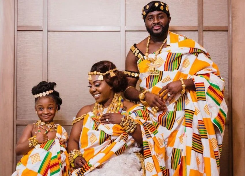 Foto de um casal e uma menina vestidos com roupa de cerimônia de casamento típica do Gana.