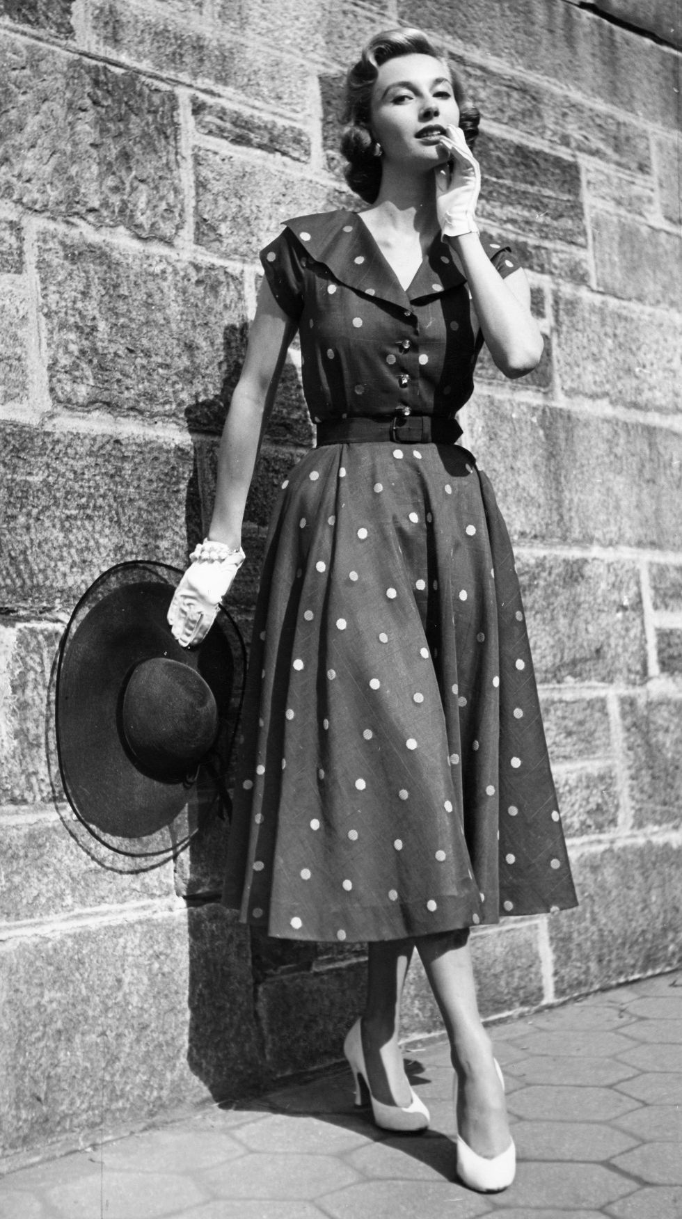 Uma modelo posando com um vestido de verão dos anos 50, com luvas brancas e chapéu preto nas mãos. A Moda e Cidadania nos anos 40 e 50.