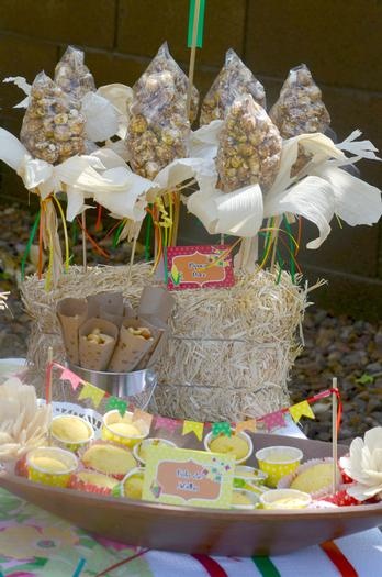 A imagem representa decorações de festa junina: pipoca doce em formato de espiga de milho enfeitada com a palha seca