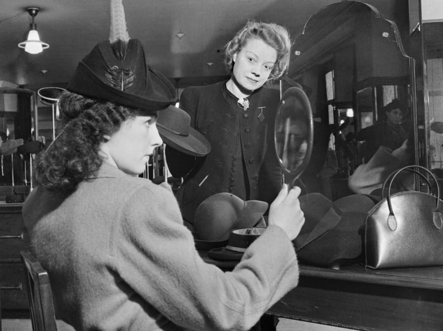 Foto em preto em branco de uma mulher provando um chapéu em uma chapelaria com um espelho na mão e sendo observada pela vendedora