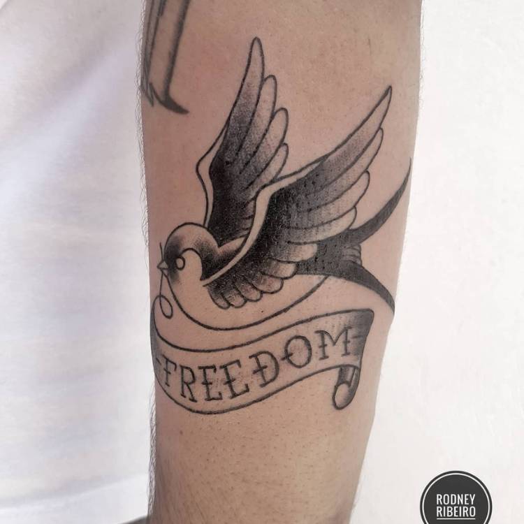 Pessoa de pele clara com tatuagem preta de andorinha old school com faixa escrito "freedom"