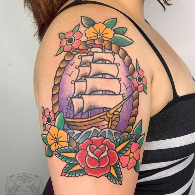 Mulher de pele clara usando regata branca e preta com tatuagem old school de barco com flores, em roxo, vermelho, azul, verde e mais