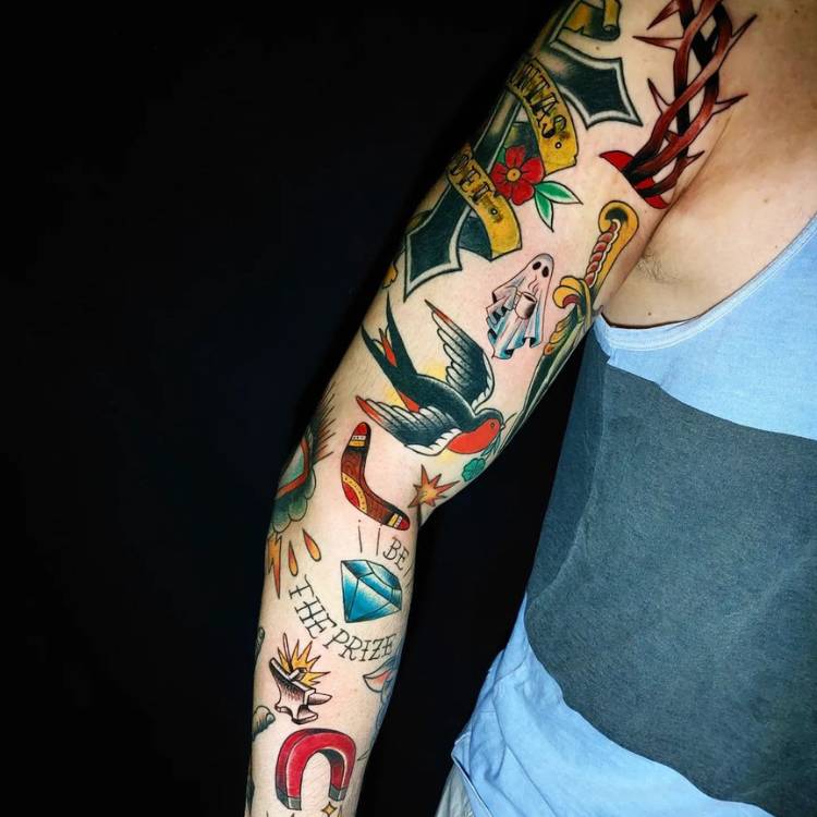 Homem de pele clara usando regata azul e braço inteiro fechado com tatuagens old school, como cruz, andorinha, adaga e mais