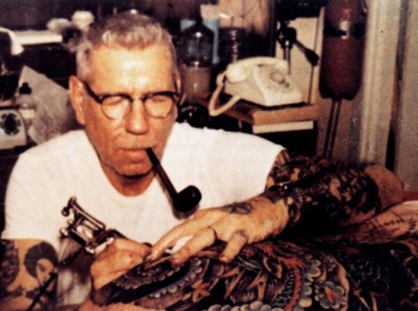 Homem de óculos, pele branca e fumando cachimbo tatuando uma pessoa