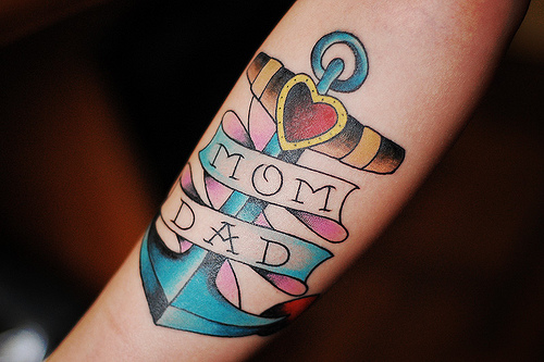 Tatuagem old school de âncora marrom, amarela e azul com faixa escrito "mom" e "dad"