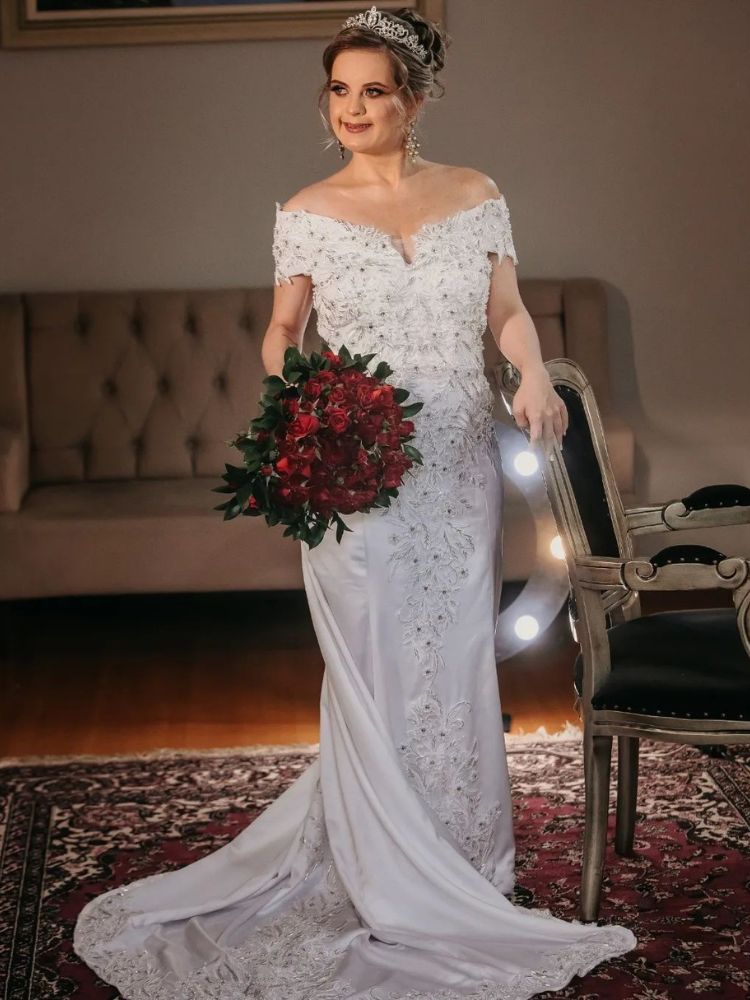 Mulher de pele clara usando vestido de noiva ombro a ombro com bordados e segurando buquê de flores vermelhas