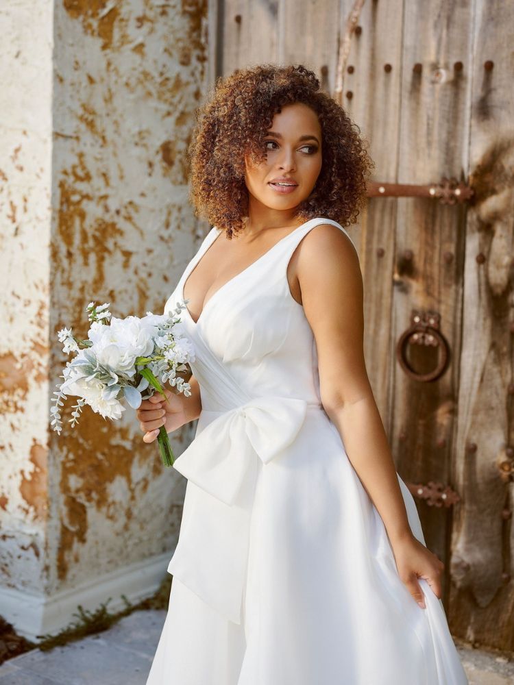 Mulher de pele negra com buquê de flores brancas usando vestido de noiva de alça com grande laço no quadril 