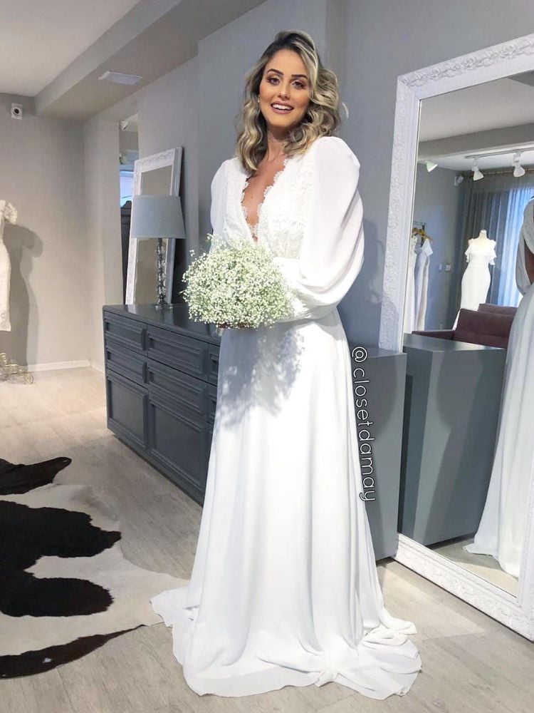 Mulher de pele clara em loja de vestidos de noiva usando vestido minimalista branco com mangas volumosas e segurando buquê de flores claras 