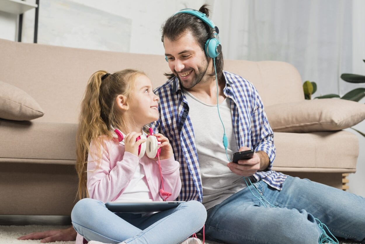 Um pai e uma filha sentam no chão da sala, ambos de headphone colorido. Ela com um Ipad, ele com um smartphone. Eles se olham e sorriem.