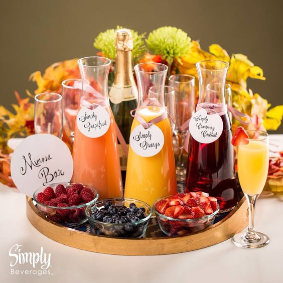 drinks em garrafas e taças de vidro, sobreposto em uma bandeja com potinhos de vidro com frutas como framboesa e morango. Ao fundo, flores