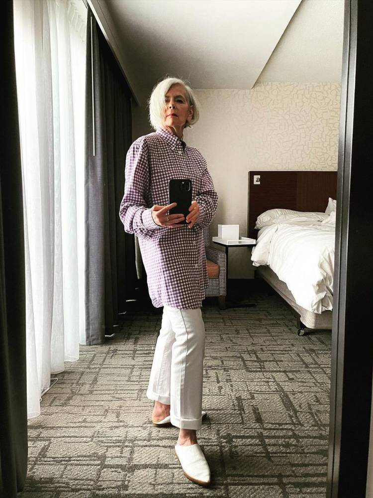 Senhora de cabelo branco tirando foto no espelho usando camisa xadrez larga e calça branca