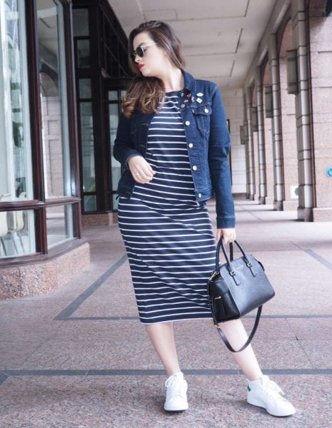 Mulher de pele branca usando vestido midi preto e branco com jaqueta jeans, bolsa preta e tênis branco