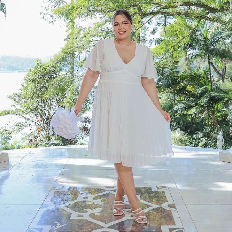 Mulher loira de pele clara usando vestido midi fluido branco e segurando buquê de flores brancas