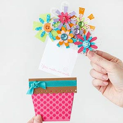 Pessoa de pele clara segurando cartão de dia das mães dentro de vaso de papel e flores coloridas de papel e botões