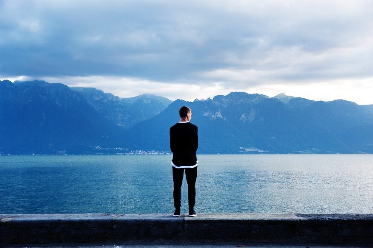 Foto de homem sozinho em paisagem de tons frios de lago com montanhas ao fundo - Como lidar com a solidao