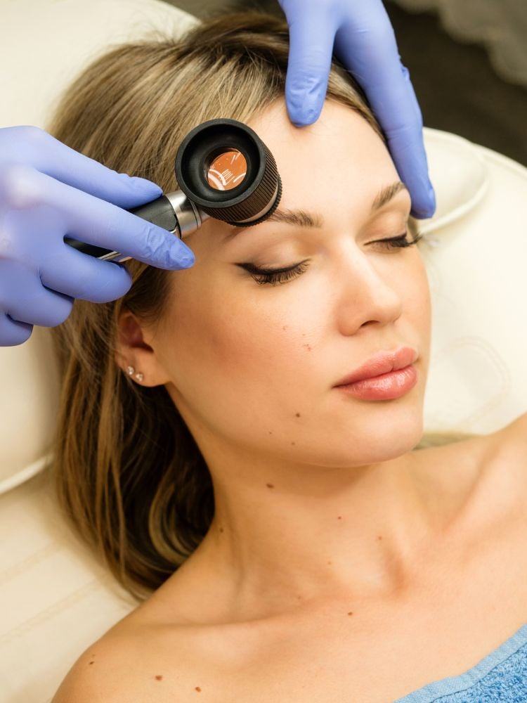 Mulher de pele clara no dermatologista, deitada enquanto uma pessoa usando luvas azuis e instrumento de ver a pele