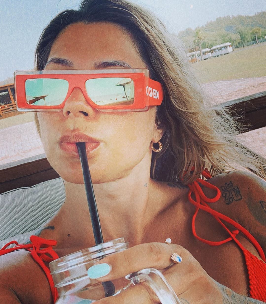 Dani Souza usando óculos de sol vermelho tomando água. 