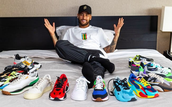 O jogador Neymar aparece deitado em sua cama com vários tênis sobre o lençol branco. Com calça jeans e camiseta branca ele está com os braços para cima em espécie de comemoração Neymar assina contrato com Puma 2