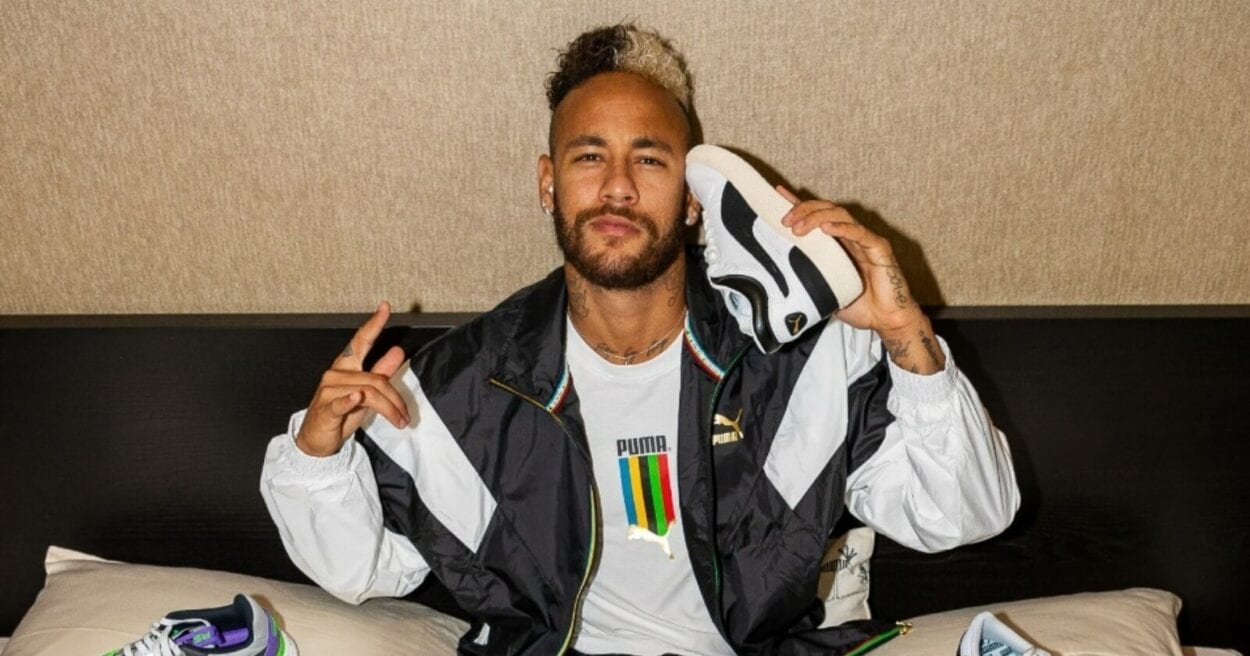 O jogador Neymar aparece deitado em sua cama com vários tênis sobre o lençol branco. Com calça jeans e camiseta branca ele está com os braços para cima em espécie de comemoração Neymar assina contrato com Puma 3
