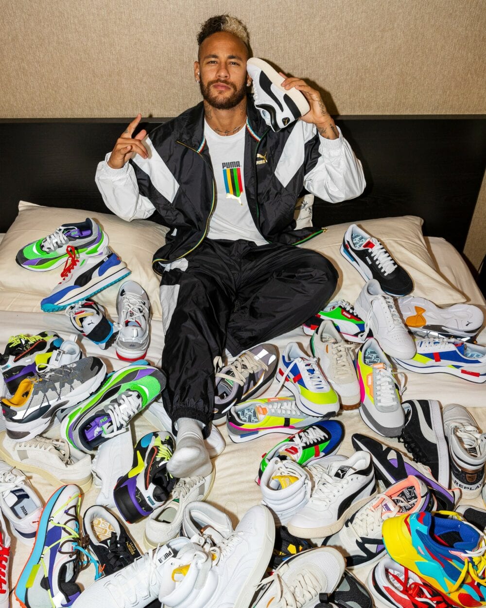 O jogador Neymar aparece deitado em sua cama com vários tênis sobre o lençol branco. Com calça jeans e camiseta branca ele está com os braços para cima em espécie de comemoração Neymar assina contrato com Puma