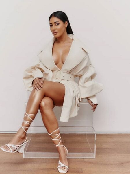 Simaria aparece sentada numa cadeira de acrílico usando trench coat nude e sandálias de tiras amarradas na canela. Com decote e cinto marcando a cintura, cabelo preto penteado para trás Simaria está curada da covid-19!