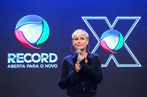 Xuxa aparece no centro da imagem usando camisa azul escura, segurando microfone e brincos de argola. Ao fundo os logos do programa dela e da Record Xuxa confirma saída da Record