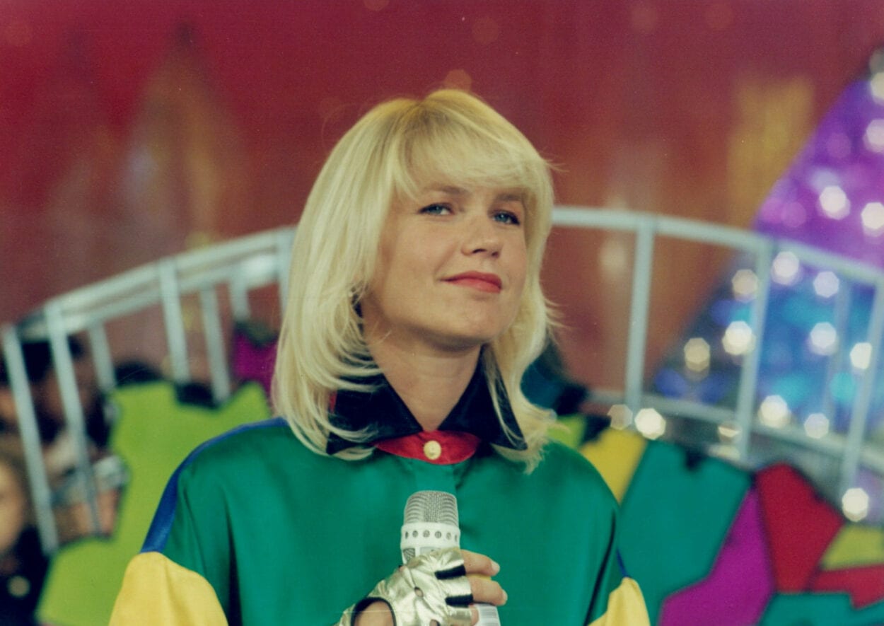 Xuxa com camisa verde clara e gola azul, ela aparece segurando um microfone com uma luva metálica. Ao fundo detalhes do cenário do antigo programa dela na Rede Globo Xuxa confirma saída da Record
