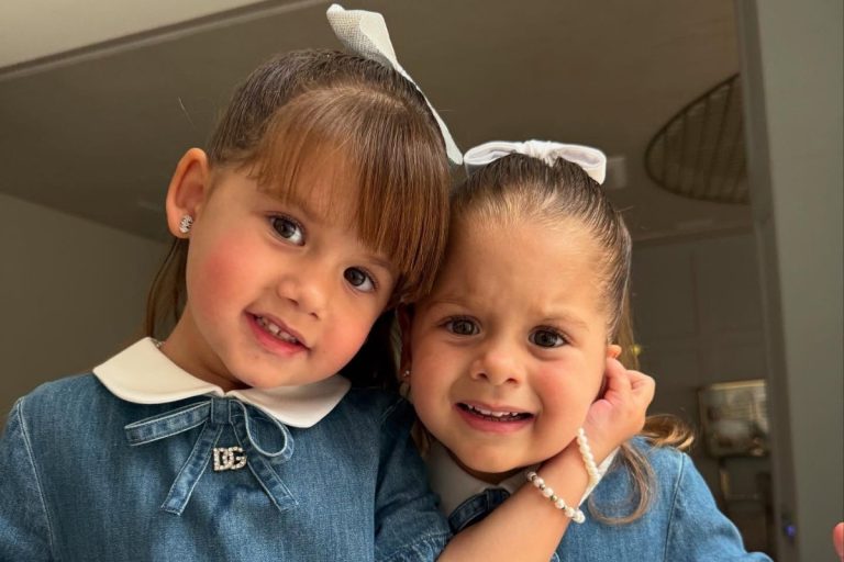 Duas meninas pequenas posam juntas, sorrindo levemente, vestindo vestidos jeans com gola branca e laços decorativos no cabelo. Uma segura carinhosamente o rosto da outra. O ambiente ao fundo é iluminado e contém móveis e elementos decorativos.