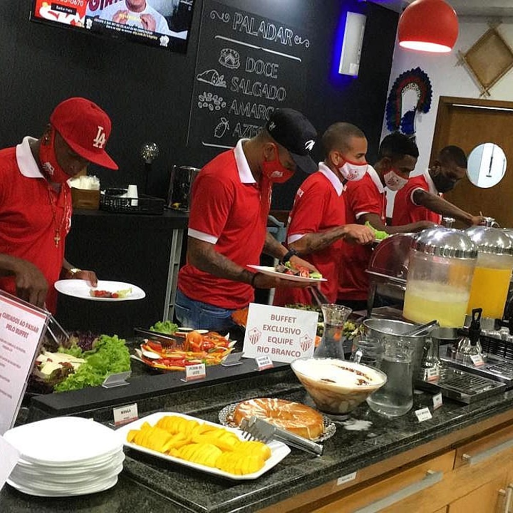 Homens de vermelho, comendo comida em buffet. Jogadores fazendo refeição em buffet exclusivo.