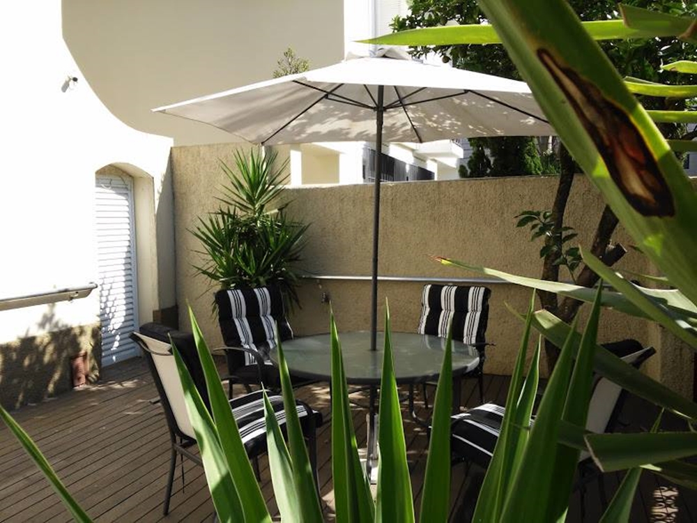 Área externa com mesa coberta para proteção do sol e cadeiras pretas e brancas. Vegetação. Ambiente externo, com cobertura e plantas