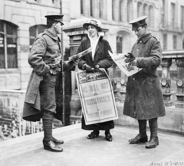 Sufragista mostrando um jornal a dois oficiais britânicos e segurando um poster que diz "Nós não vamos ser prussianizados". 1915. 