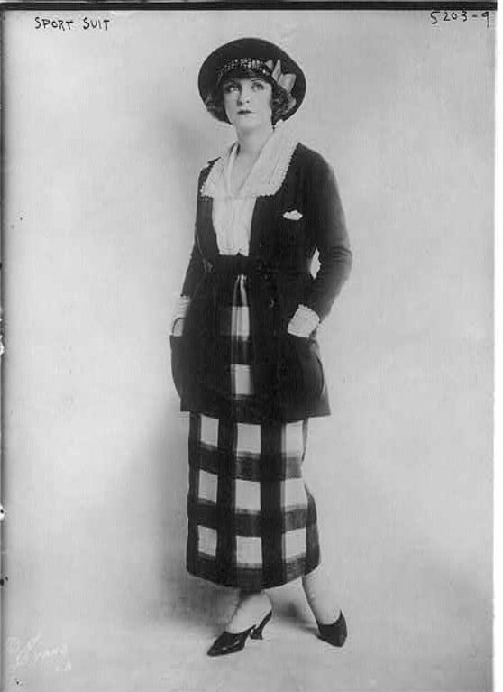 Mulher posando com um modelo de roupa dos anos 1920, com uma saia longa xadrez e chapéu. Look da Moda e Cidadania nos anos 20 e 30.
