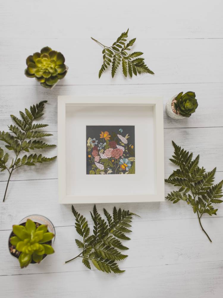 Imagem de um quadro de flores com moldura branca rodeado por folhagens e vasos de suculenta. 