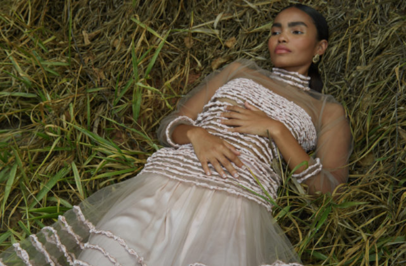 Modelo deitada na grama usa vestido transparente com aplicação de listras bordadas na gola, punho, barra e torso. 