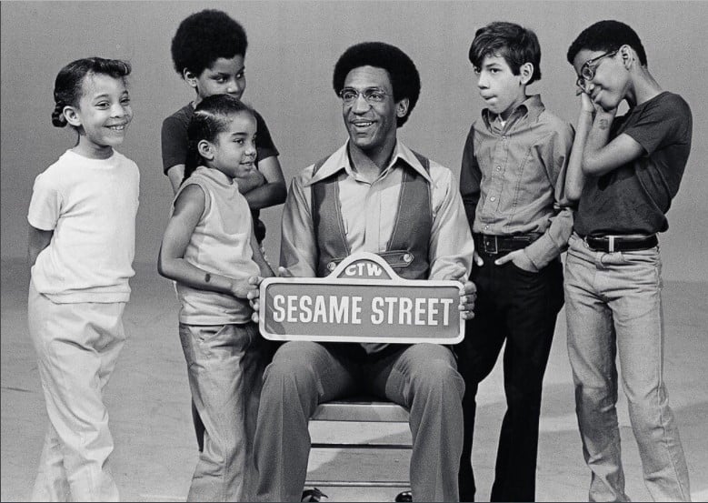Fotos dos anos 70 de um grupo de crianças ao redor de Bill Cosby a segurar uma placa da Vila Sésamo.