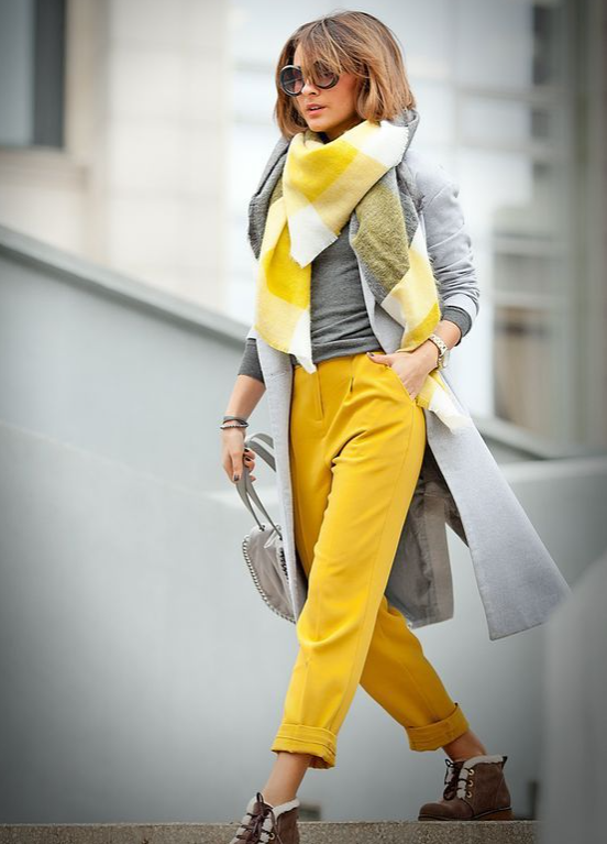 Mulher usa calça amarela, sobretudo e blusa cinza e cachecol xadrez