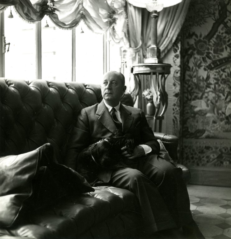 Foto em preto e branco de Christian Dior com o seu cachorro no seu apartamento, em Paris, em 1950. Christian Dior com o seu cachorro no seu apartamento, em Paris, em 1950.