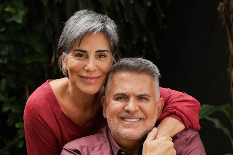 Um casal maduro e sorridente posa em um ambiente ao ar livre, com a mulher abraçando o homem por trás. Ambos vestem roupas em tons avermelhados, transmitindo afeto e harmonia. Plantas esverdeadas compõem o fundo natural da imagem.