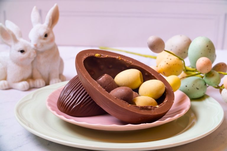 Metade de um ovo de chocolate recheado com miniovos coloridos está sobre um prato rosa e verde pastel. Ao fundo, dois coelhos decorativos brancos e ovos decorativos em tons suaves completam o cenário delicado de Páscoa.