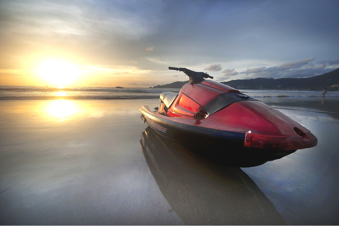 Imagem de um Jet Ski preto e vermelho estacionado em uma praia durante o pôr do sol.