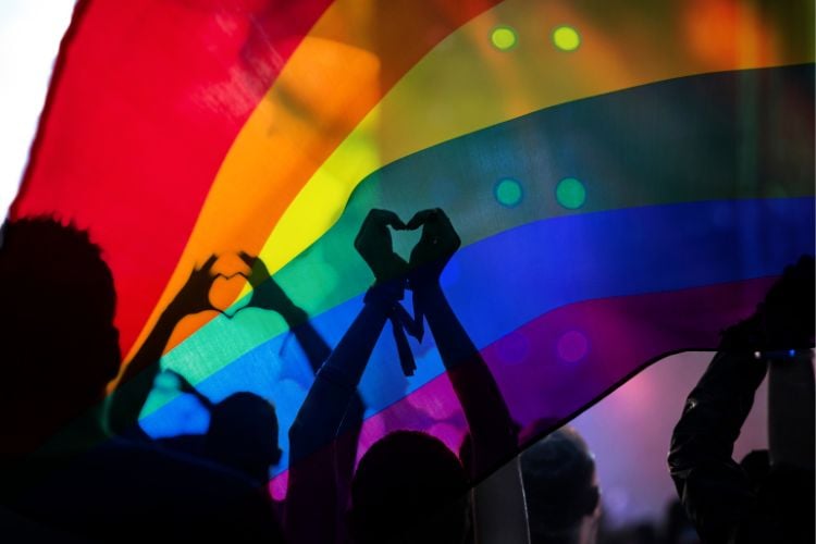 Pessoas sob enorme bandeira LGBT colorida, sendo duas delas fazendo coração com as mãos