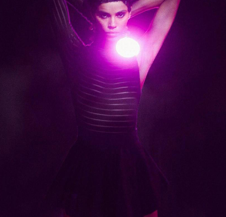 Modelo usando um vestido preto, com uma luz forte e roxa atrás dela.