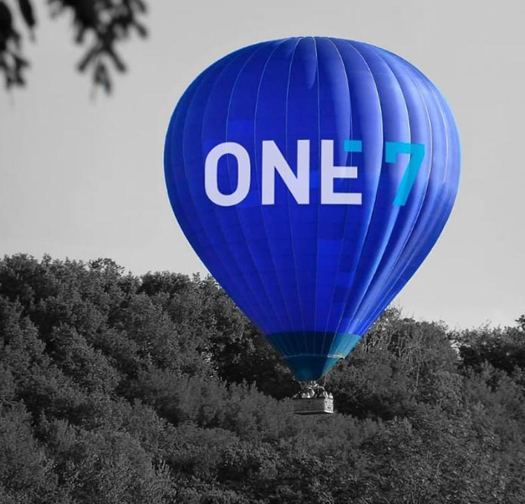Fundo da imagem é uma floresta em preto e branco. Um balão azul com One7 escrito está no centro da foto.